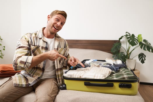 Handsome smiling man, pointing at his suitcase with clothes, invites for holiday, encourages to pack and go on vacation, sits on bed with luggage photo