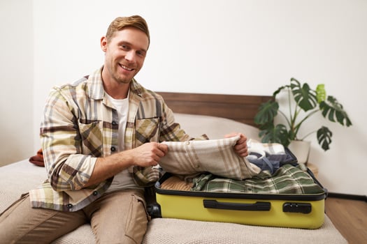 Portrait of young man sits on bed with suitcase, goes on holiday, packs clothes for vacation or business trip, checks his luggage photo