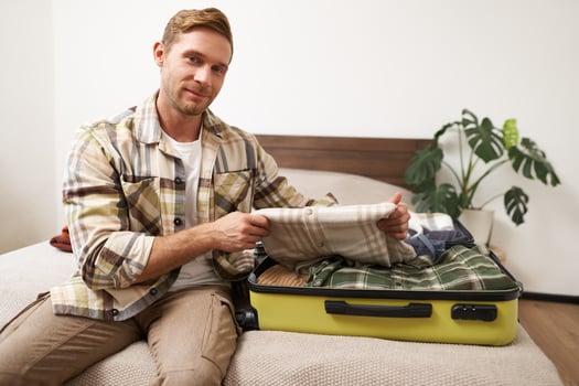 Portrait of man, tourist in hotel, packing or unpacking his suitcase, holding shirt, taking clothes out of luggage bag, smiling at camera photo