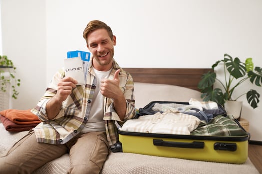 Portrait of happy, excited young man, tourist holds his passport and plane tickets, packs clothes in suitcase, gets ready for summer holiday, goes on vacation and feels excitement photo