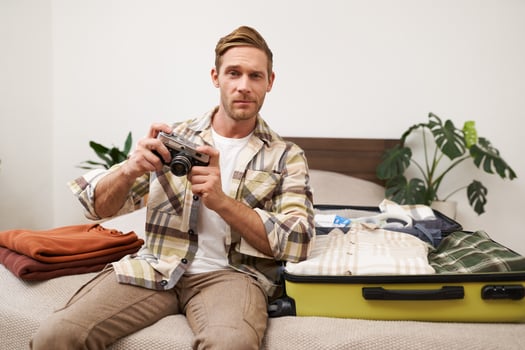 Portrait of handsome tourist, male model holding a camera, sitting near opened suitcase with clothes, man going on vacation, packing for summer holiday retreat photo