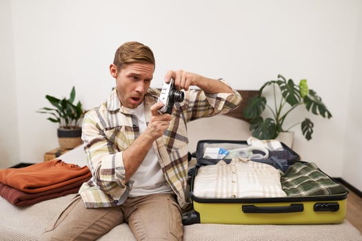 Image of young man, tourist checks his camera, sits on bed with opened suitcase, photographing, unpacking clothes on holiday, going on vacation photo
