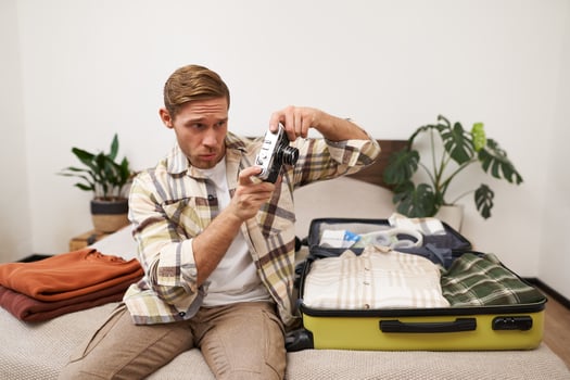 Portrait of young man sitting on bed, looking through camera, takes a picture, sits and packs clothes in suitcase, goes on vacation, tourist gets ready for holiday, summer retreat photo