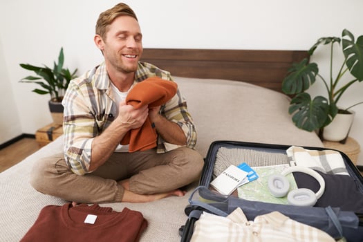Portrait of smiling young man sits on bed with suitcase, packing for summer holiday, smelling clean and fresh clothes, goes on vacation photo