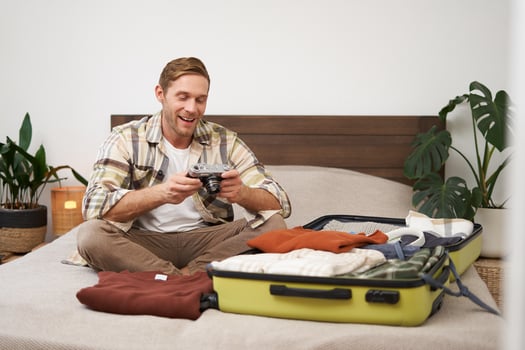 Portrait of handsome, happy young man, sitting with suitcase on bed, looking at his camera, laughing and smiling, watching photos or s from holiday trip