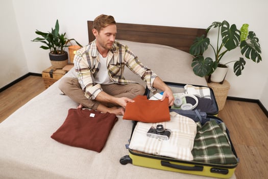 Portrait of young handsome man, tourist packing his clothes in suitcase, going on vacation, summer trip, prepares luggage for holiday, sits on bed in room photo