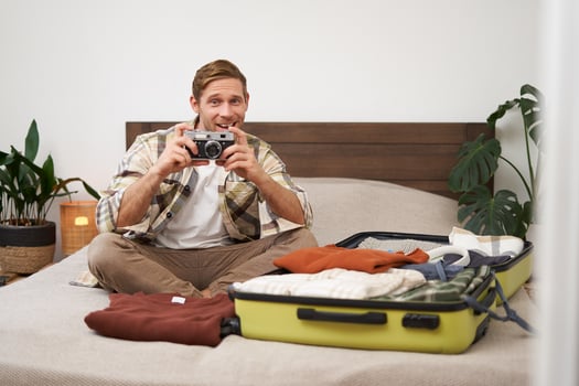 Cheerful young man takes a picture on digital camera, sits on bed, goes on holiday, packs clothes for vacation, tourist excited before going on trip, travelling photo