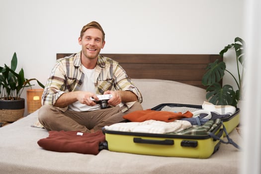 Portrait of young excited man, sits on bed with suitcase, going on holiday, holding digital camera, going through photo album on gadget, preparing for summer vacation