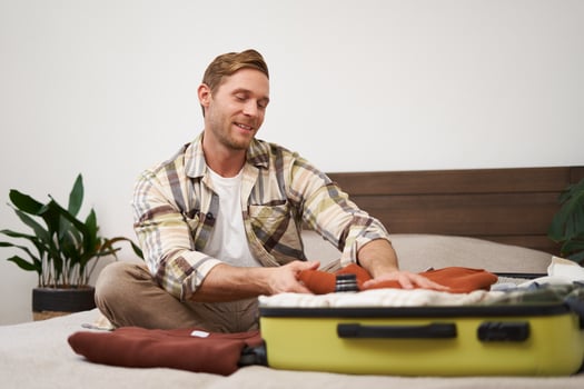 Portrait of smiling handsome man, sits on bed, puts clothes inside suitcase, goes on vacation, dreams about holiday, books a trip with travel agency and packs things photo