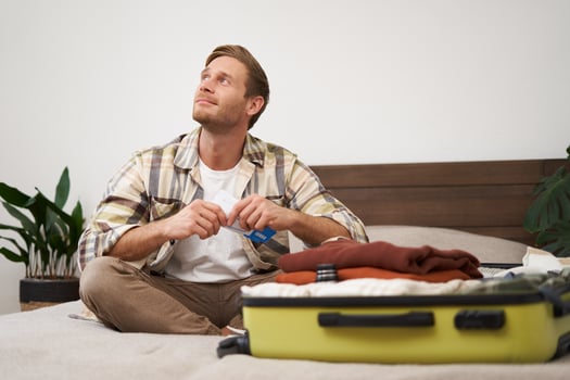 Portrait of handsome man, tourist, looking at his holiday tickets, booked vacation with travel agency, sitting on bed and packing clothes in suitcase photo