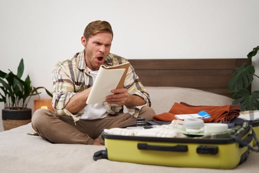 Portrait of angry man, packing suitcase, looking frustrated at notebook with check list of items for holiday, preparing to go on vacation photo
