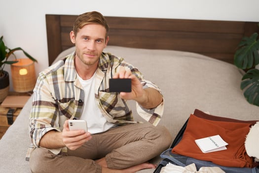 Portrait of tourist, man packing suitcase with clothes and passport, holding mobile phone, showing his credit card photo