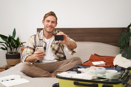Portrait of happy man buying tickets with credit card cashback, sitting on bed and packing clothes for holiday, putting items in suitcase, holding mobile phone photo