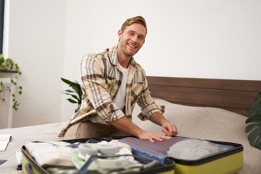 Portrait of young handsome bearded guy, tourist going on holiday, fastening his clothes in suitcase, packing for vacation, going on business trip, sitting in bedroom photo