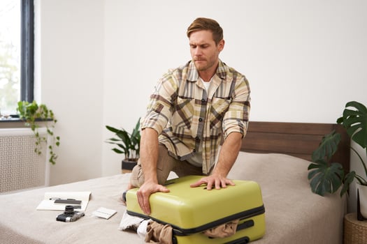 Portrait of young man, tourist packs his bag, closing full suitcase, trying to push the luggage inside to zip the bag, sitting on bed in bedroom photo