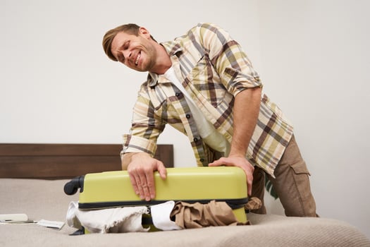 Image of young man, tourist struggling to close his suitcase full of clothes, pushing on bag, trying to press items inside luggage to shut the zipper, packing for travelling abroad, going on holiday photo