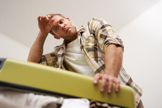 Image of tired strong man, trying to close his suitcase with clothes, sweating, wiping forehead after putting an effort to pack luggage for business trip or a holiday photo