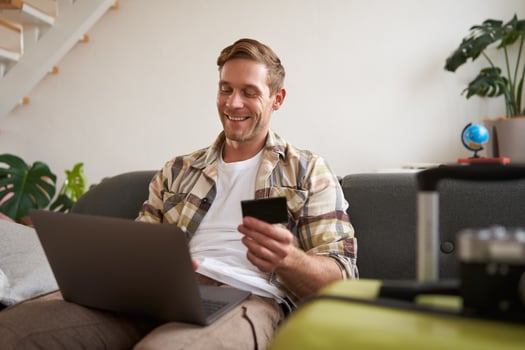 Happy man booking holiday online on laptop, paying for vacation on travel agency website, buying flight tickets in internet, sitting in living room with suitcase photo