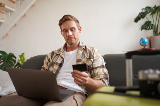Portrait of cheerful guy buying flight tickets online, booking hotel by using laptop and credit card, sitting near suitcase in living room photo