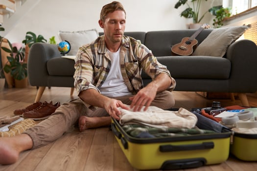 Image of young man, traveller, sitting on floor with opened suitcase, packing clothes for a trip, going on holiday photo