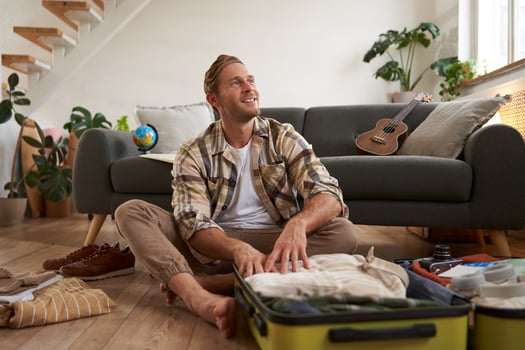 Happy young traveller, man sits with luggage on floor, looking aside and smiling, thinking about his future holiday, going on vacation, packing suitcase photo