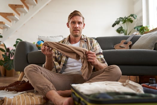 Portrait of confused man, sitting in living room with suitcase, holding pair of trousers, looking clueless, struggling to pack for holiday photo