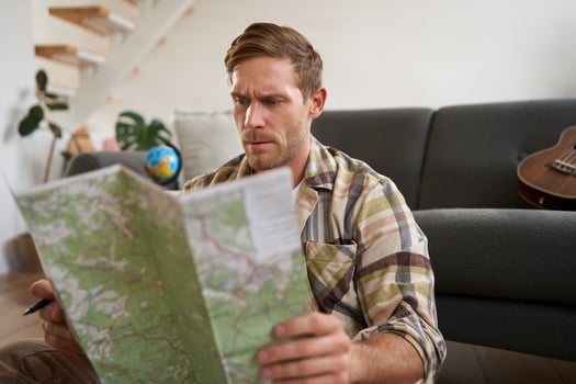 Portrait of happy young man, tourist with suitcase, sits in living room and looks at map, chooses holiday destination, picks route for travelling photo