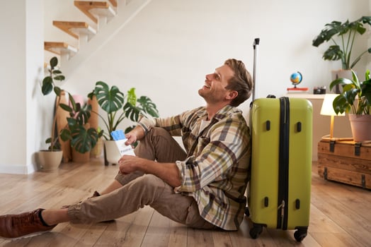 Portrait of male model with suitcase, sitting on floor, holding flight tickets and passport, going abroad, waiting for a taxi to airport photo