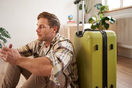 Portrait of young man, traveller sits with suitcase and mobile phone, chats on smartphone photo