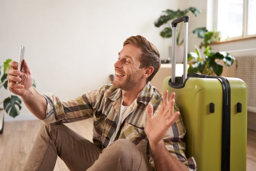 Portrait of happy tourist, man takes selfies before his flight, photos with suitcase, uses smartphone to post pictures on social media