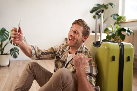 Cheerful smiling man with suitcase, chats using smartphone, takes selfie, goes on holiday photo