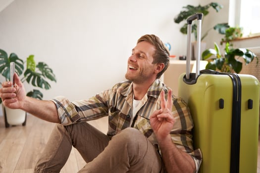 Portrait of smiling handsome man, shows peace sign, takes selfies on smartphone, sits with suitcase, goes on holiday photo
