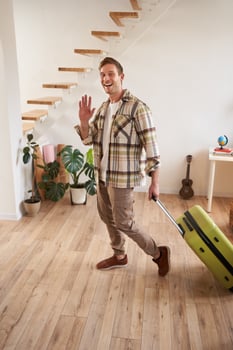 Vertical shot of happy young man with suitcase, checking in the rented apartment, waving hand at camera, walking with luggage indoors photo