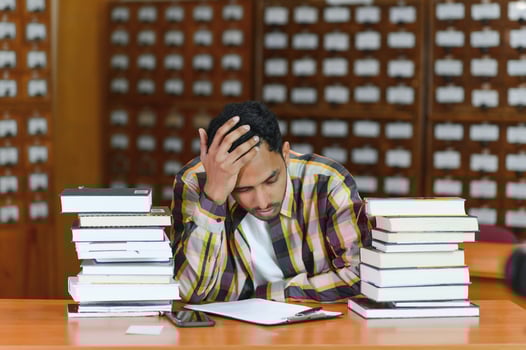 Portrait of cheerful male international Indian student with backpack, learning accessories standing near bookshelves at university library or book store during break between lessons. Education concept photo