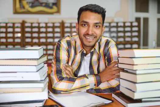 Portrait of cheerful male international Indian student with backpack, learning accessories standing near bookshelves at university library or book store during break between lessons. Education concept photo