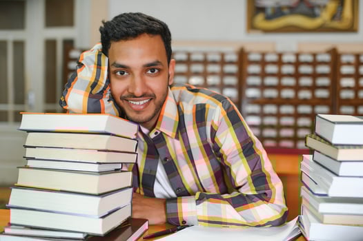 Portrait of cheerful male international Indian student with backpack, learning accessories standing near bookshelves at university library or book store during break between lessons. Education concept photo