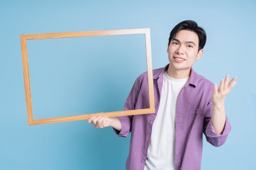 Young Asian man holding photo frame on blue background