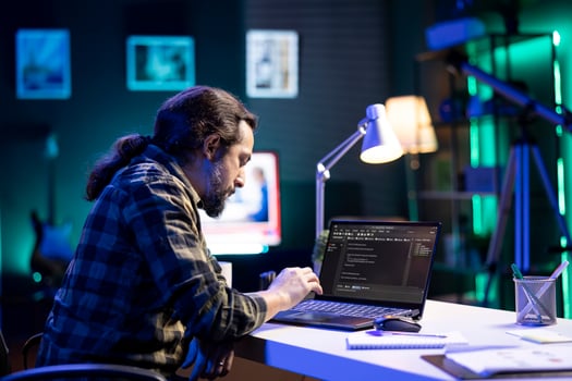 Dedicated male programmer uses his portable computer to manage databases and keep an eye on network security. Bearded man is coding on a digital laptop while seated at a desk. photo