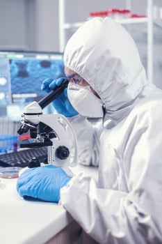 Researcher in modern microbiology laboratory uses microscope dressed in ppe suit. Virolog in coverall during coronavirus outbreak conducting healthcare scientific analysis. photo