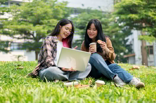 Two young Asian female college students are doing homework on a laptop together, sitting on grass. photo