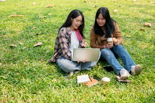 Two young Asian female college students are doing homework on a laptop together, sitting on grass. photo