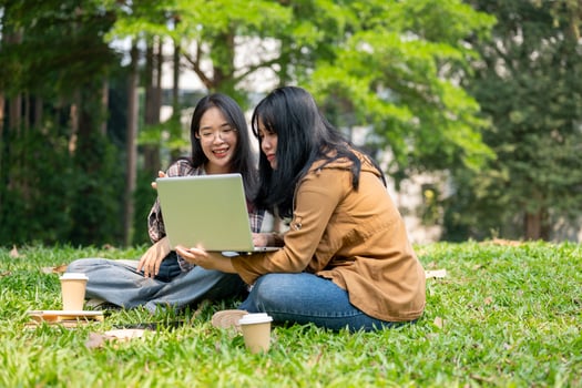 Two young Asian female students are sitting on the grass in a park, studying together with a laptop. photo