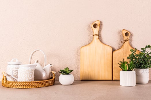 White ceramic tea set on round wicker tray among indoor plants on kitchen countertop. Kitchen background in light colors. A copy space. Front view photo
