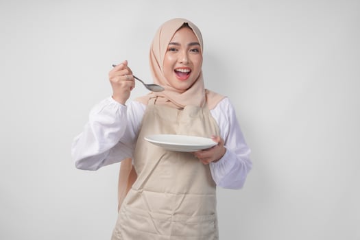Hungry young Asian Muslim woman in hijab and cream apron holding spoon and eating from an empty plate with copy space over it. Ramadan concept photo