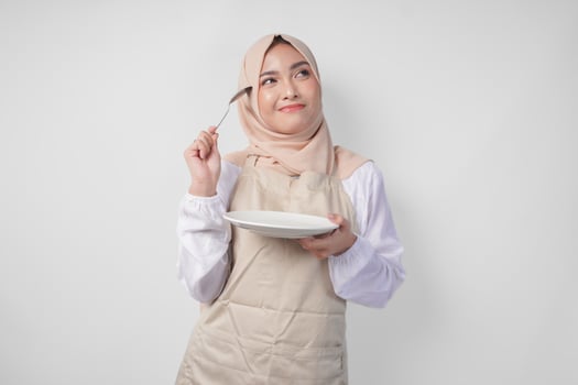 Confused young Asian Muslim woman in hijab and cream apron holding spoon and empty plate with copy space over it, thinking hard what food menu to eat for iftar. Ramadan concept photo