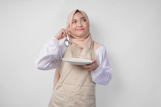 Confused young Asian Muslim woman in hijab and cream apron holding spoon and empty plate with copy space over it, thinking hard what food menu to eat for iftar. Ramadan concept photo