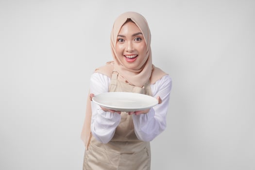 Beautiful young Asian Muslim woman in hijab and cream apron presenting an empty plate with copy space over it, showing the food menu for iftar photo