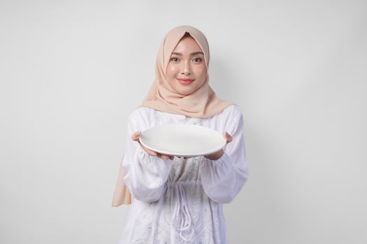 Beautiful young Asian Muslim woman in hijab presenting an empty plate with copy space over it, showing the food menu for iftar photo