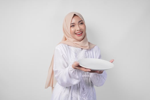 Beautiful young Asian Muslim woman in hijab presenting an empty plate with copy space over it, showing the food menu for iftar photo