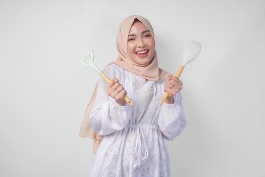 Portrait of a happy Asian Muslim woman in veil hijab holding spatula and kitchen cooking utensils while smiling cheerfully. Ramadan concept photo